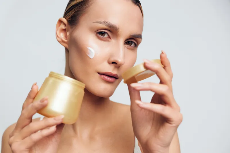 Woman skincare moisturizer cream beauty jar portrait, holding a golden cream jar and applying moisturizer to her face, studio close up, clean skin, calm wellness mood