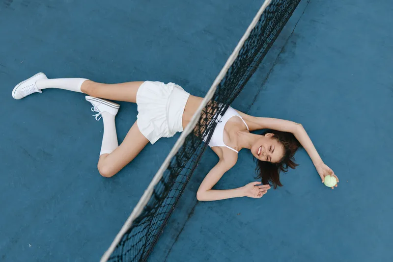 A young woman in sporty attire lounging on a blue tennis court, exuding joy and playfulness while holding a tennis ball, showcasing a vibrant and fresh summer vibe