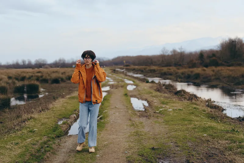 Woman lifestyle outdoors walking river path nature jacket, orange tones on scarf and coat, blue jeans, calm countryside fields, overcast daylight, natural colors, relaxed confident stance, breathing