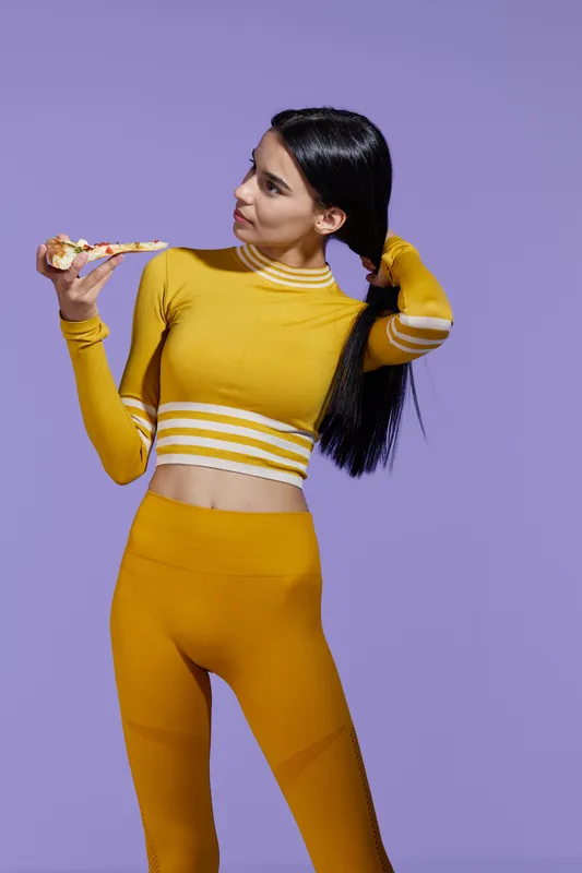 Sport fitness woman in yellow activewear eating slice of pizza, showing balance of healthy lifestyle and indulgence, with confident posture on purple background.