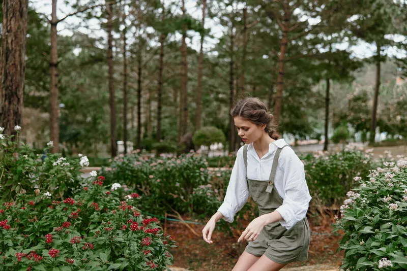 Young woman in a garden wearing a white shirt and green overalls, enjoying nature surrounded by blooming flowers, evoking feelings of serenity and joy