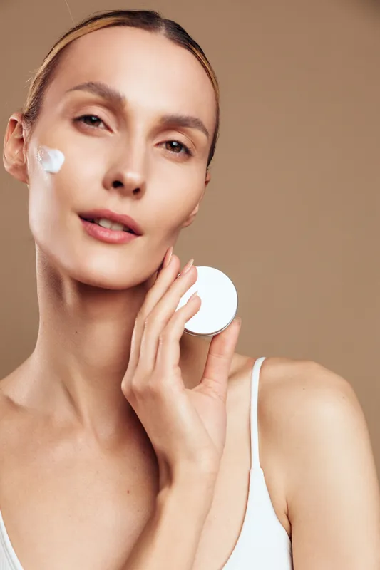Young woman with clear skin applies face cream on her cheek while holding a cosmetic pad against a neutral beige background with a calm expression.