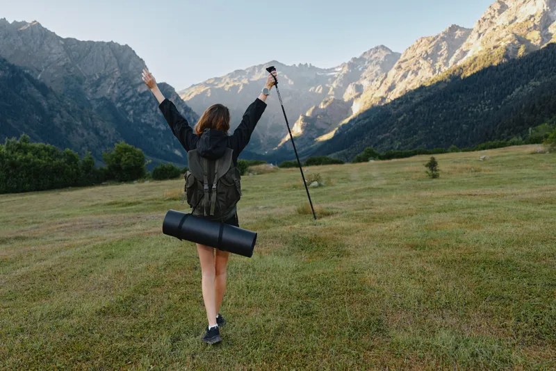 A lone hiker stands in a grassy valley between towering peaks, arms raised in triumph. The traveler wears a backpack and gazes toward the distant mountains, savoring a moment of adventure and