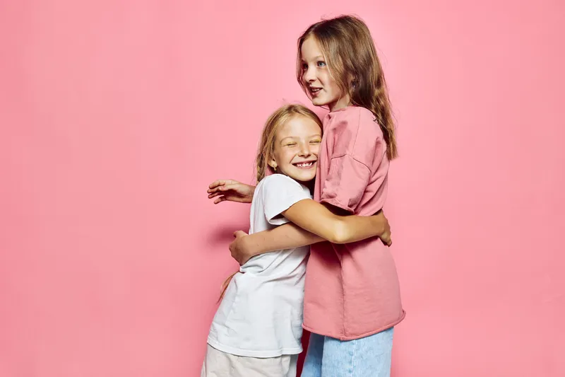 Two happy young girls hugging each other affectionately in front of a pink background. They smile warmly showing friendship and sibling love. Studio portrait.
