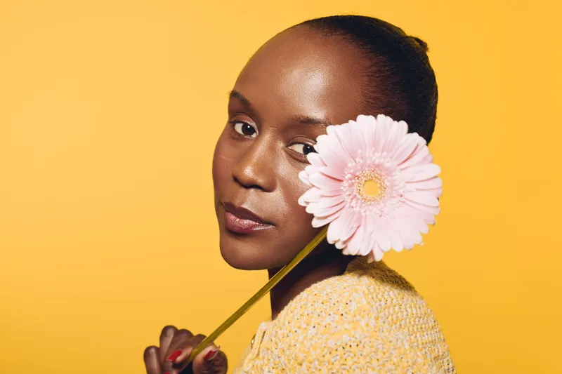 Close-up portrait of dark skinned African woman holding pink flower near face against yellow background. Concept for beauty, skincare, nature, and natural cosmetics with vibrant colors and soft light.