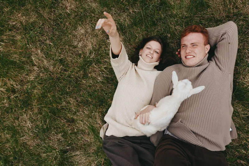 Cheerful couple outdoors lying on grass, taking a selfie together, warm sweaters, relaxed mood, sunny day, friendly dog nestled between them, natural colors, casual lifestyle moment, candid and joyful