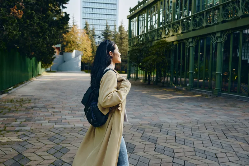 Woman with backpack walks along an urban street, wearing a trench coat, enjoying a calm outdoor moment in a city setting with glass buildings and trees, conveying fashion, travel, and everyday