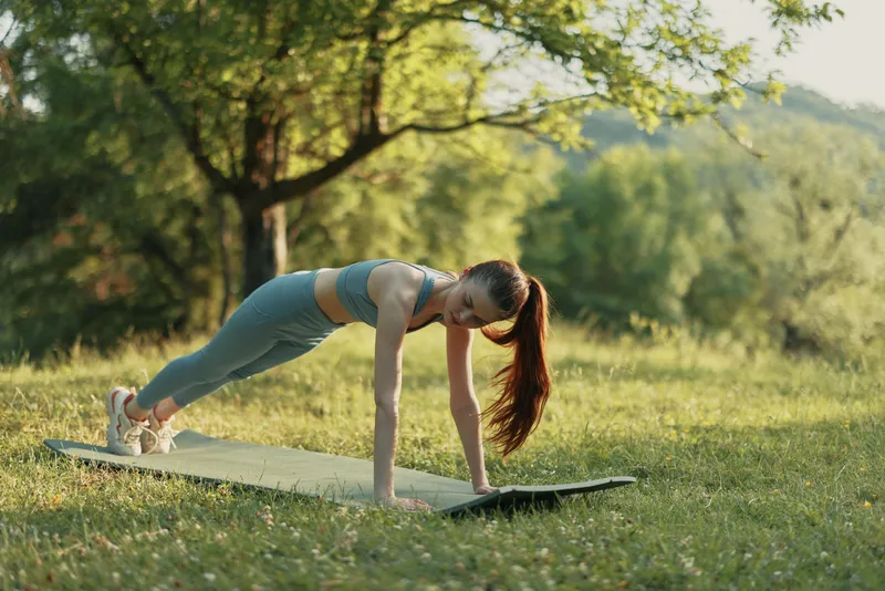 Young woman performing outdoor exercise in a plank position on a yoga mat, showcasing fitness and wellness in a sunny green park setting