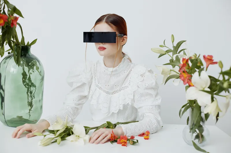 A futuristic model with sleek sunglasses poses elegantly with white and colorful flowers in a minimalist setting, highlighting beauty, fashion, and innovation