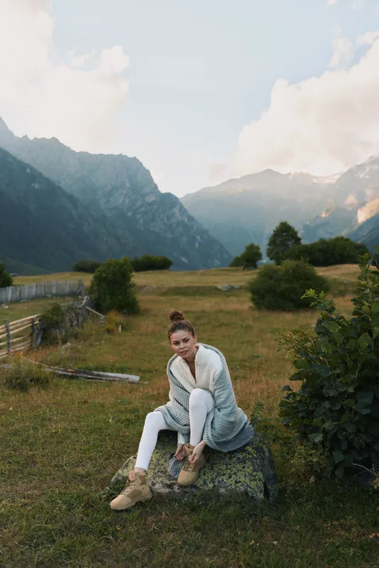 Woman lifestyle in nature, sitting on rock, tying boots, casual outfit, mountain valley background, peaceful atmosphere, soft natural light, outdoor activity