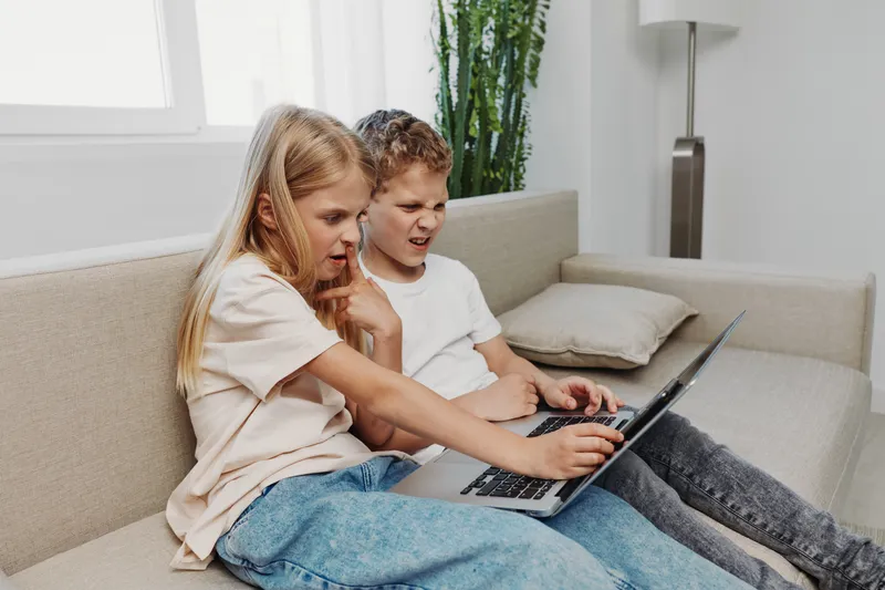 Curious children sitting on beige sofa using laptop together, focused on screen in bright living room with modern decor and natural light.