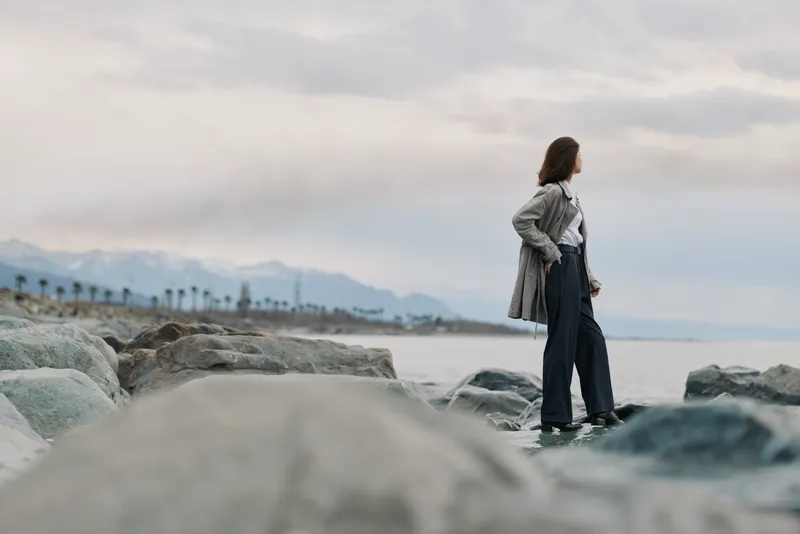 Woman in casual outfit stands on rocky shore looking at sea during cloudy day. Calm coastal landscape with distant mountains and palm trees in background. Concept of contemplation and nature.