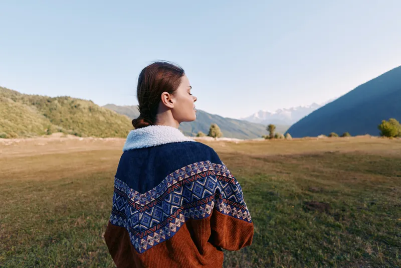 Woman in patterned sweater stands in a mountain landscape, overlooking a wide meadow and clear sky. Outdoor portrait conveys calm nature scene, travel and peaceful solitude.