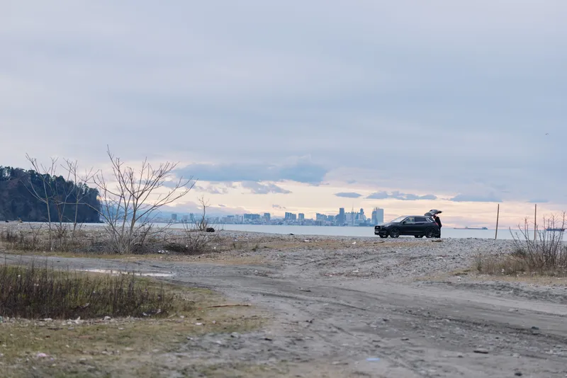 Car on a dirt road beside a lake with distant city skyline and cloudy sky, a rugged travel mood and sense of solitude by the water
