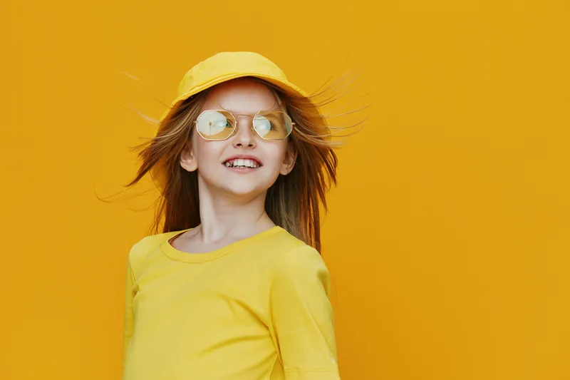 Cheerful girl wearing a yellow outfit and cap, posing against a vibrant yellow background, embodying joy and summer spirit with sunglasses on