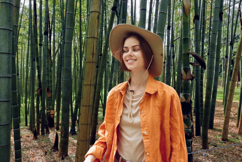 Woman walking in a bamboo forest wearing a hat and orange jacket, smile and portrait in nature. Outdoors travel and leisure scene among green trees conveying calm exploration.
