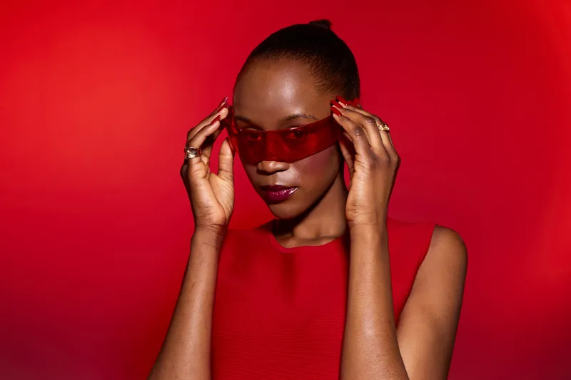 Elegant black African woman with dark skin wears red sleeveless top and red sunglasses, posing confidently against a vivid red background. Concept of fashion, style, and beauty.