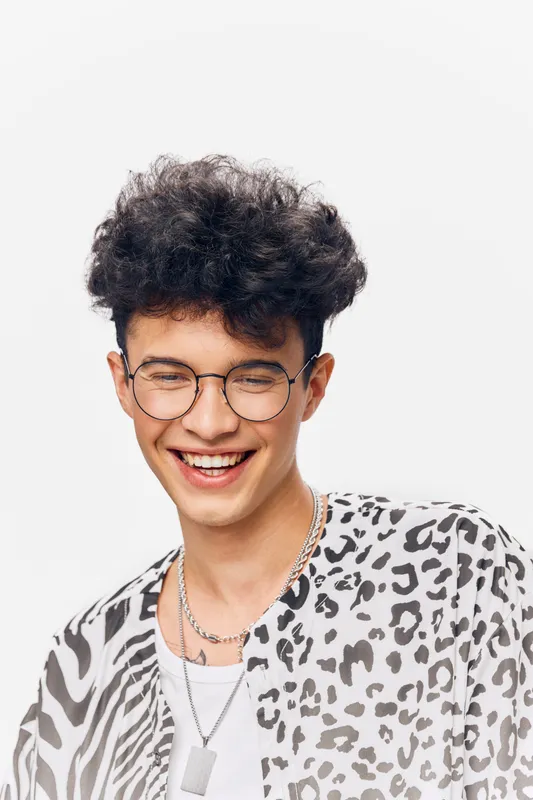 Smiling man with curly hair wearing glasses and a trendy animal print shirt, conveying happiness and youthfulness in a bright studio setting.