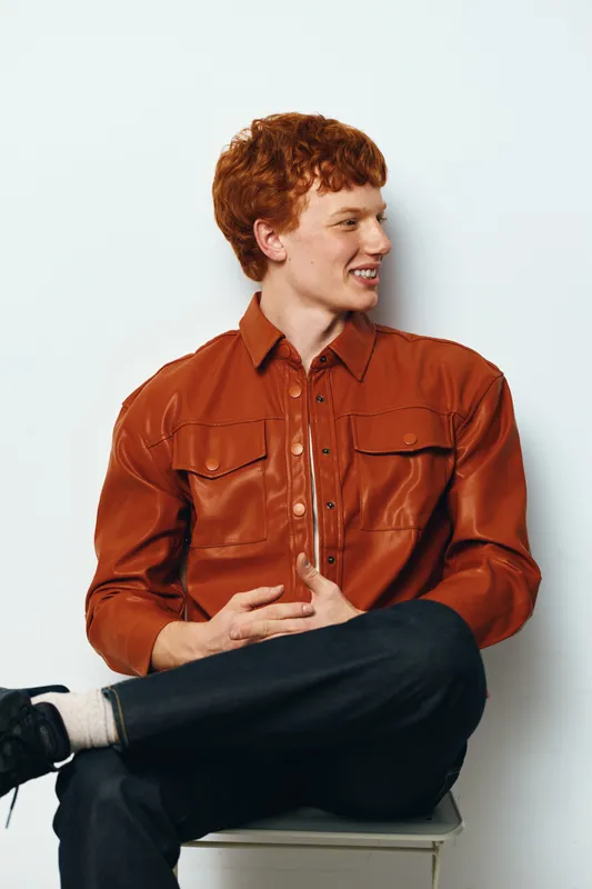 Young man with red hair in orange leather jacket sitting on stool looking sideways with smile on white background. Casual fashion, confident pose and relaxed mood indoors.