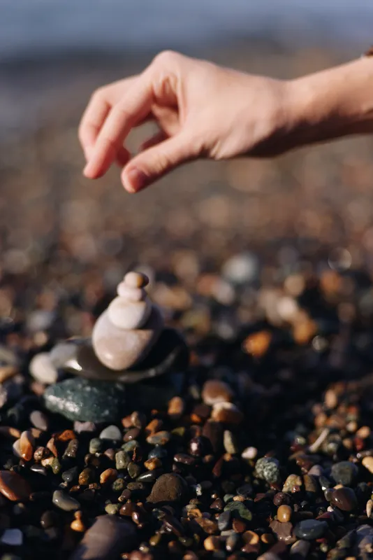 Close up of a hand stacking smooth stones on a pebble beach, forming a tiny zen cairn amid coastal rocks and waves, conveying calm balance and focus