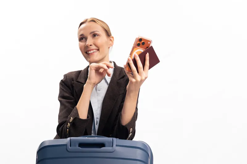 Smiling young woman with suitcase holds smartphone and passport cover, ready for travel. White background studio image conveys concepts of vacation, trip planning, and tourism.