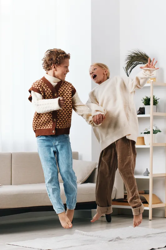 Two adults dance hand in hand in a bright living room, wearing cozy knitwear and relaxed jeans, capturing a candid, joyful moment and warm home energy.