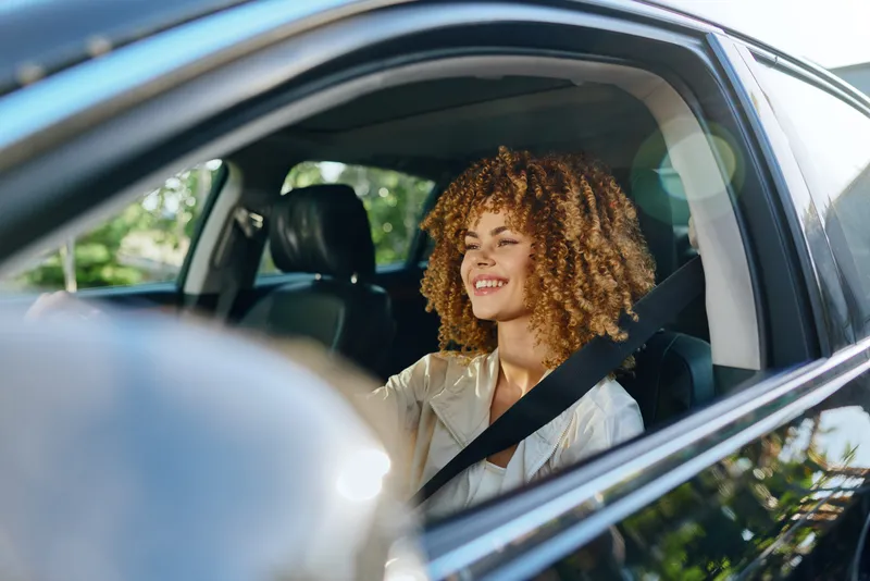 Smiling woman driving car fastened with seatbelt, curly hair, casual clothing, daylight, safe urban travel, joyful expression, natural light.