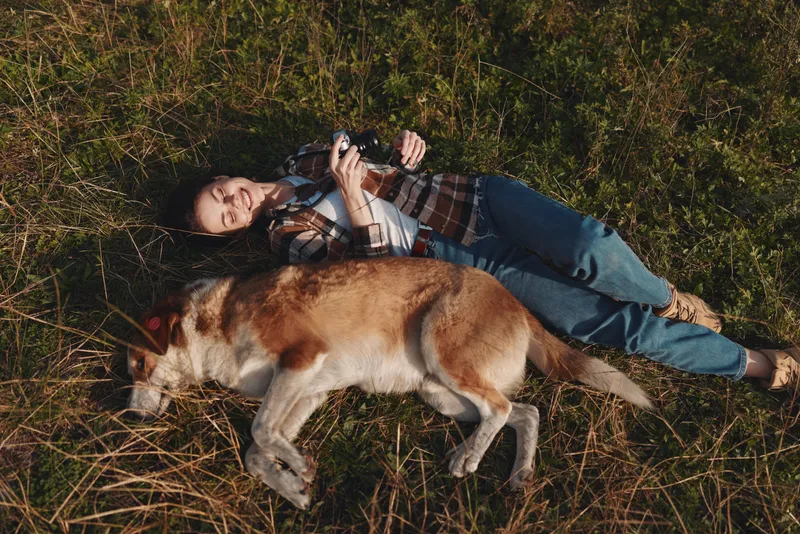 Woman lying on grass with a dog beside, outdoor relaxation and companionship, casual clothing, sunlit field, peaceful moment for rest, nature inspired bond and calm.