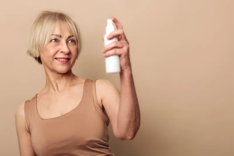 Mature woman with short blonde hair sprays facial mist on her skin, wearing beige tank top. Concept for skincare routine, hydration, anti aging, wellness, and beauty care at home.