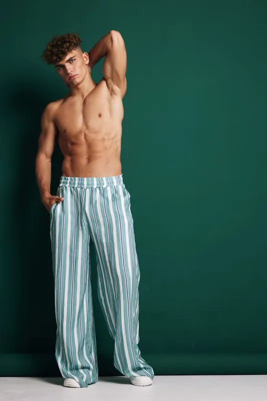 Young muscular man posing shirtless with hand behind head wearing loose striped pants and white sneakers against green background in studio setting.