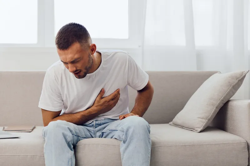 Young man sitting on the couch, experiencing discomfort in the chest area. The serene interior and neutral tones create a thoughtful atmosphere for emotional wellbeing.