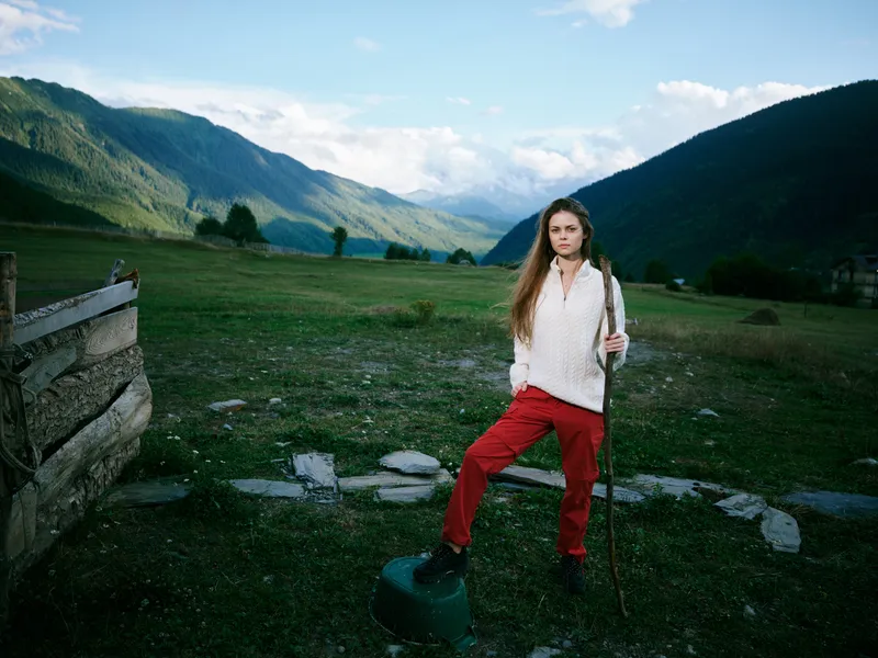 Young hiker stands with a walking stick in a green valley, framed by rolling mountains, blue sky, and open countryside, evoking serene outdoor adventure and exploration