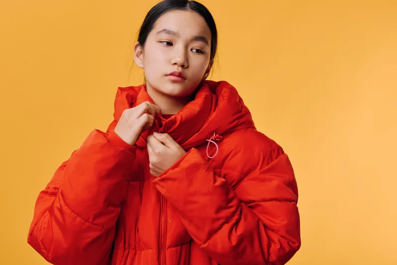 Young woman adjusting red puffer jacket with hood on orange background, fashion portrait with casual warm clothing and confident expression for winter or autumn style.