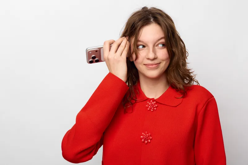 Young woman in red sweater holds smartphone near ear smiling and looking aside. Casual lifestyle concept with communication, technology, and positive emotions on white background.