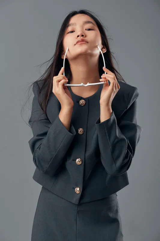 Young woman in dark suit holding sunglasses near face posing with confidence on gray background. Fashion model with long hair wearing professional clothing and modern accessories indoors.