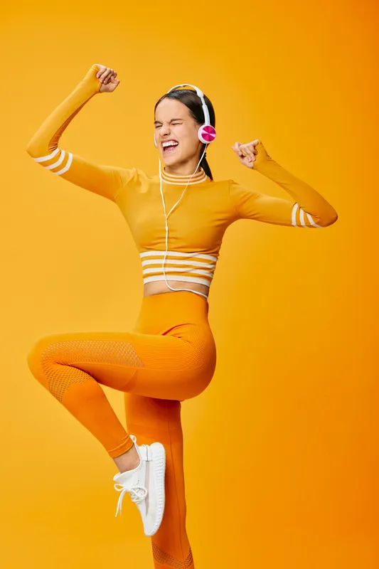 Energetic woman in orange athletic wear with headphones, jumping mid air with fists raised against a vibrant orange backdrop, illustrating fitness, sport, movement and upbeat lifestyle.