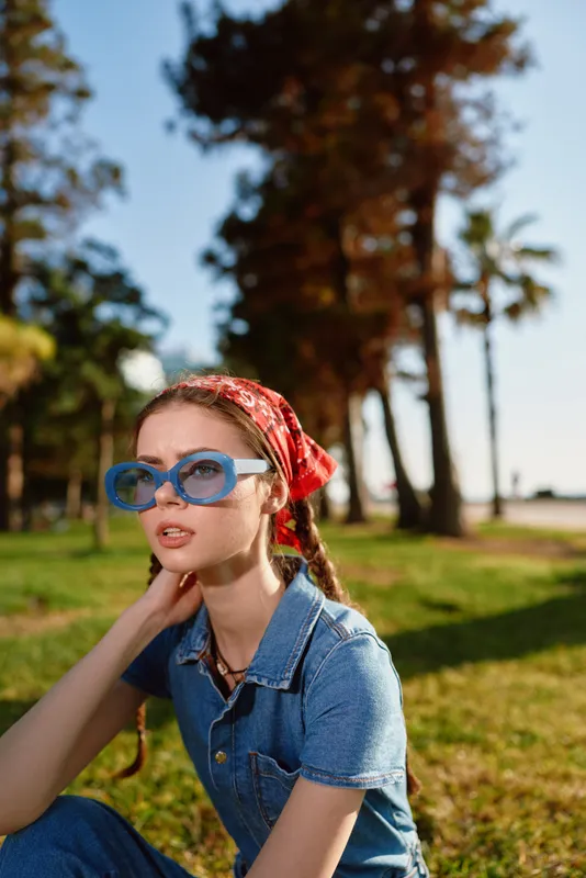Young woman with red headscarf and blue sunglasses sitting outdoors in sunny park with tall trees and green grass in warm summer light