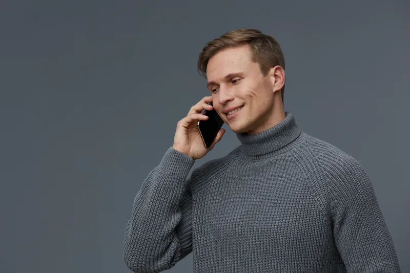 Smiling man with short brown hair wearing a cozy gray turtleneck sweater relaxedly talking on mobile phone against plain gray background studio portrait. People lifestyle concept