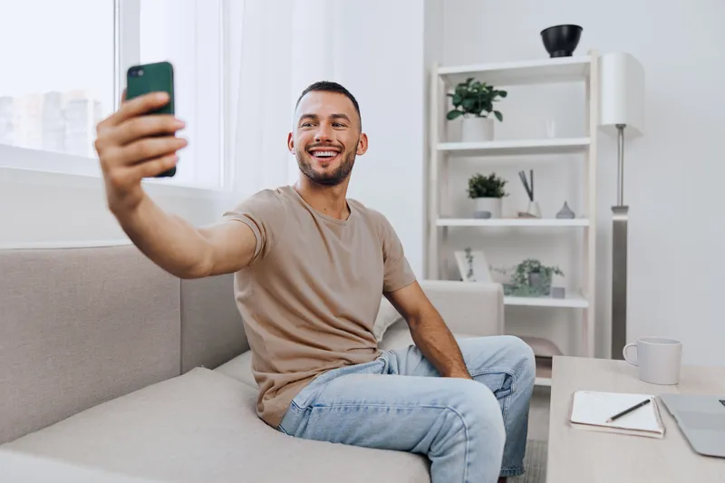 Smiling man capturing a moment on his phone, reflecting the joy of solo living and soft masculinity in a cozy home setting, promoting emotional well-being.