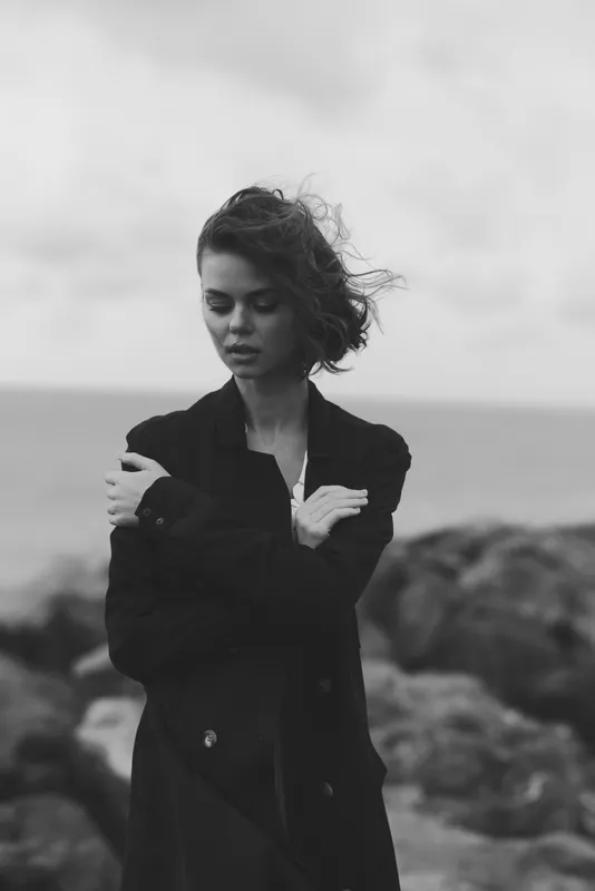 Woman in a long coat on a windy coastline, arms crossed, moody monochrome scene by the sea, showcasing fashion and solitary atmosphere.