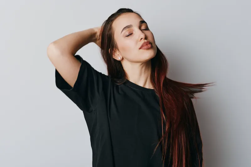 Woman with long hair and closed eyes posing with hand behind head wearing black t shirt on plain background. Female model with relaxed expression and casual style in studio.