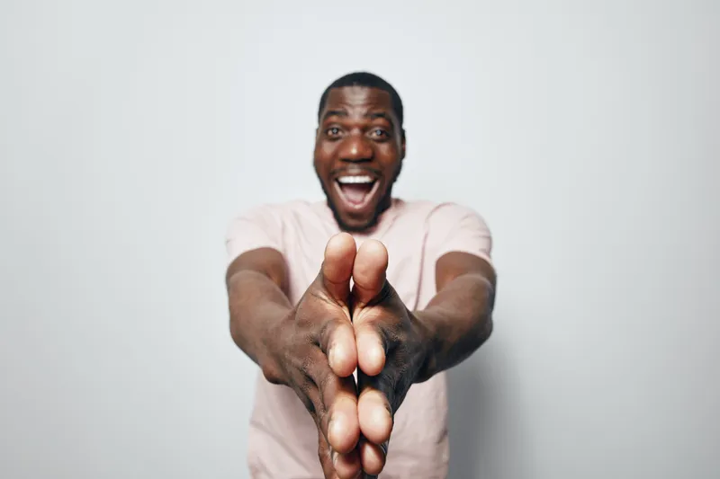 African man with dark skin wearing light shirt smiling and clapping hands together in front of white background. Happy male portrait with joyful expression and casual style.