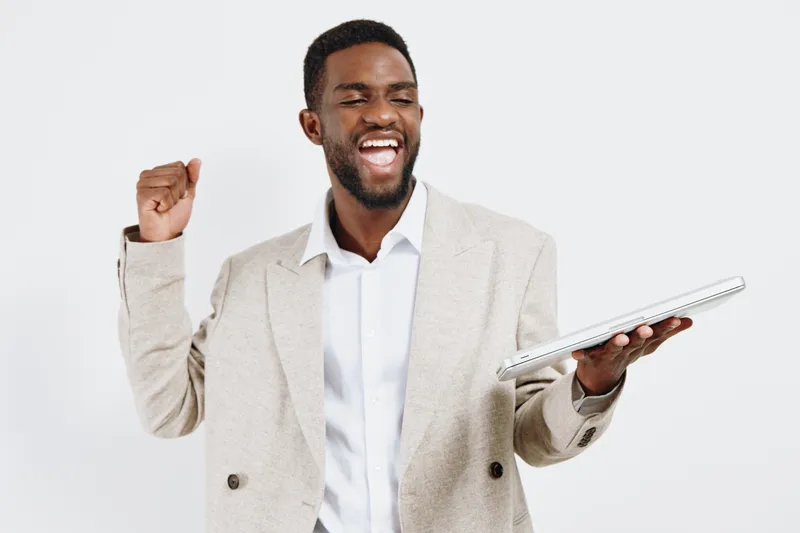 Confident man celebrating with joyful expression, wearing light beige blazer and white shirt, holding tablet in hand, isolated on plain white background, studio portrait. Business lifestyle concept.