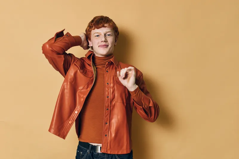 Young man with red hair in orange leather jacket and turtleneck posing confidently with hand on head on beige background. Casual style, fashion and portrait concept with happy expression.
