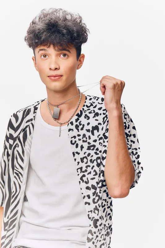 Young man with mixed animal print shirt and curly hair, holding necklace, posing confidently against white background with casual modern style and relaxed expression.