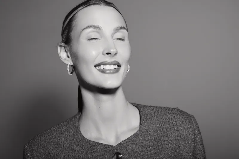 Closeup of smiling Caucasian woman with fair skin and closed eyes, wearing earrings and textured top, studio portrait with soft lighting.