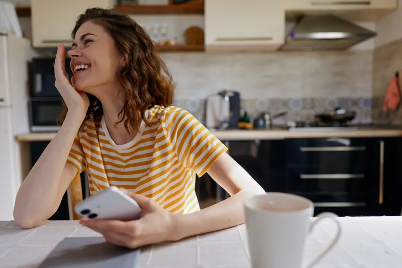 Woman lifestyle home in a bright kitchen, smiling at her smartphone, wearing a striped shirt, leaning on a table, warm light creates a casual candid moment of daily life