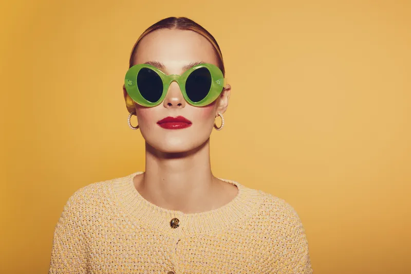 Stylish woman with slicked back hair wears large green sunglasses and red lipstick against yellow background. Fashion portrait with bold accessories and modern makeup look.