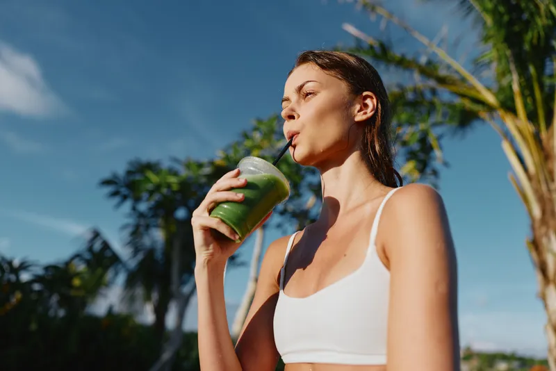 Young woman in white sports bra drinking green smoothie outdoors in sunny tropical environment. Healthy lifestyle concept with fresh juice and natural background.