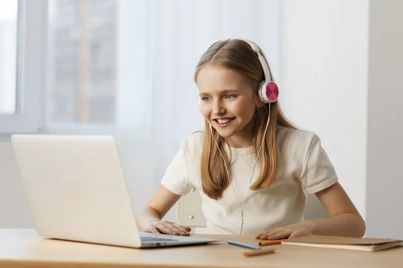 Happy young girl wearing headphones, enjoying online learning while sitting at a desk, with a bright and modern interior background